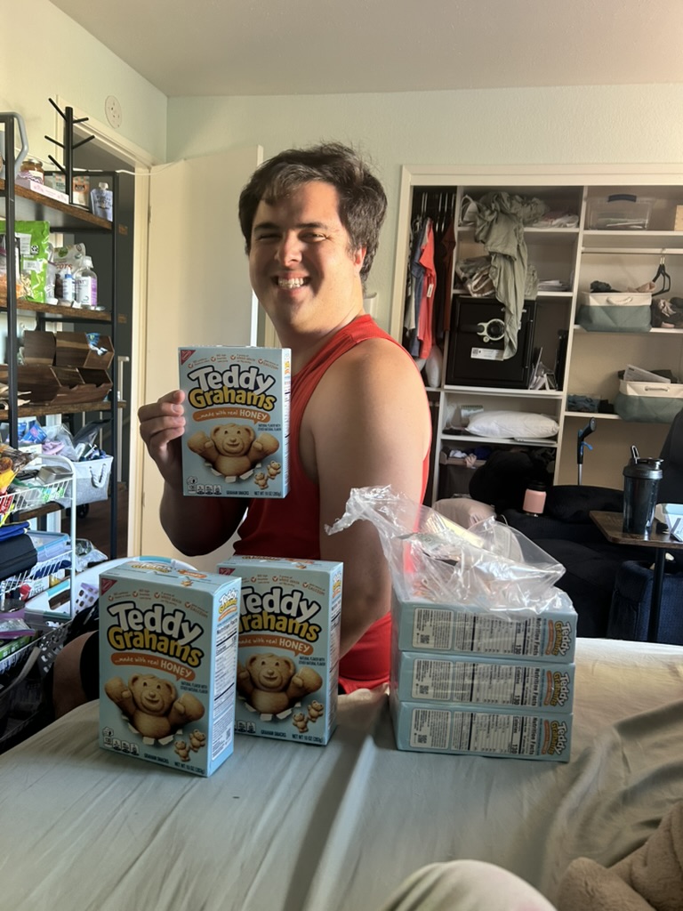 Nick smiling holding a box of Teddy Grahams while sitting on the bed with a large stack of Teddy Grahams snack boxes gathered together.