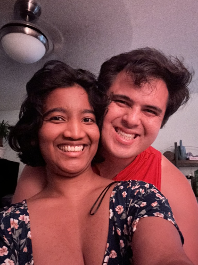A close selfie of Harshini and Nick together indoors at night, both smiling softly, with dim lighting and a calm, intimate atmosphere.