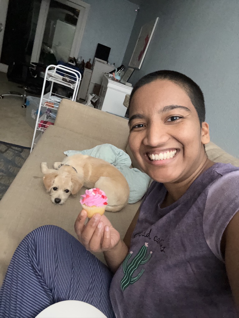 Harshini smiling while holding a pink cupcake at home after celebrating a personal milestone, with her dog Toffee resting nearby.