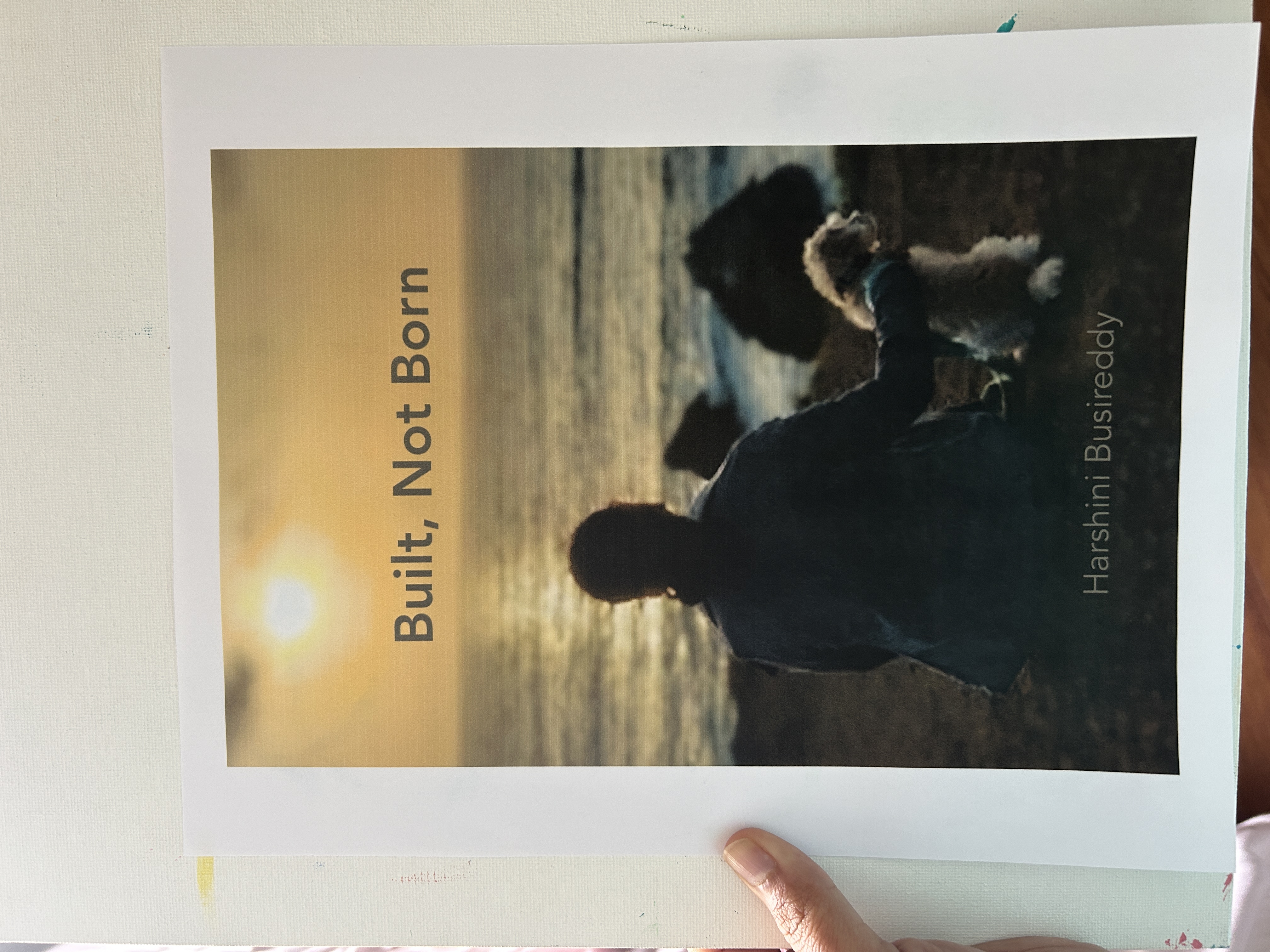 A hand holds a printed book cover titled “Built, Not Born,” showing a person sitting beside a dog facing the ocean at sunset, with the author name Harshini Busireddy at the bottom.