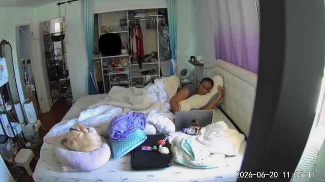 Harshini sits partially upright in bed with a laptop nearby, surrounded by blankets and pillows. The room shows an open closet, cooling equipment, and everyday living items around the bed.