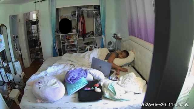 Harshini lies turned on her side in bed with a laptop close by. Pillows, blankets, and bedside items are arranged around her as part of a supported resting and working setup.