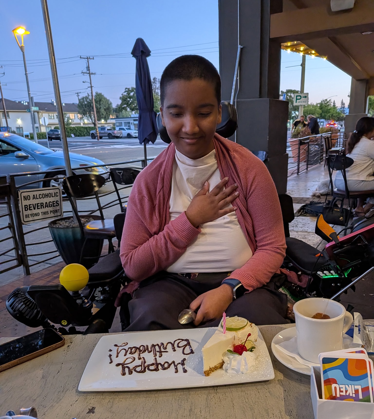 Harshini sits at a restaurant table with one hand over her heart beside a lit birthday candle and cheesecake.