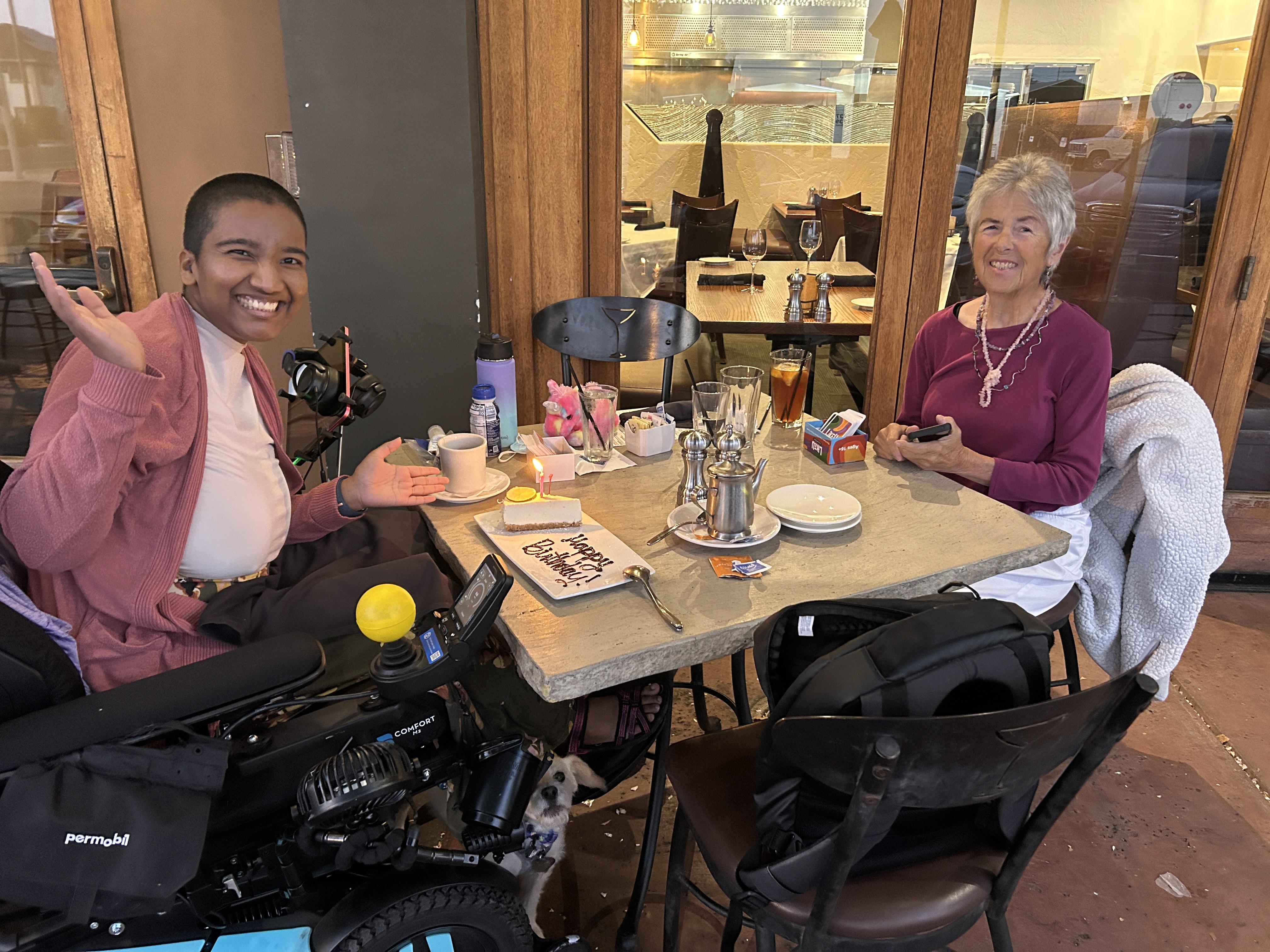 Harshini and her neighbor smile at an outdoor restaurant table with birthday cheesecake between them. Toffee is beneath the table.