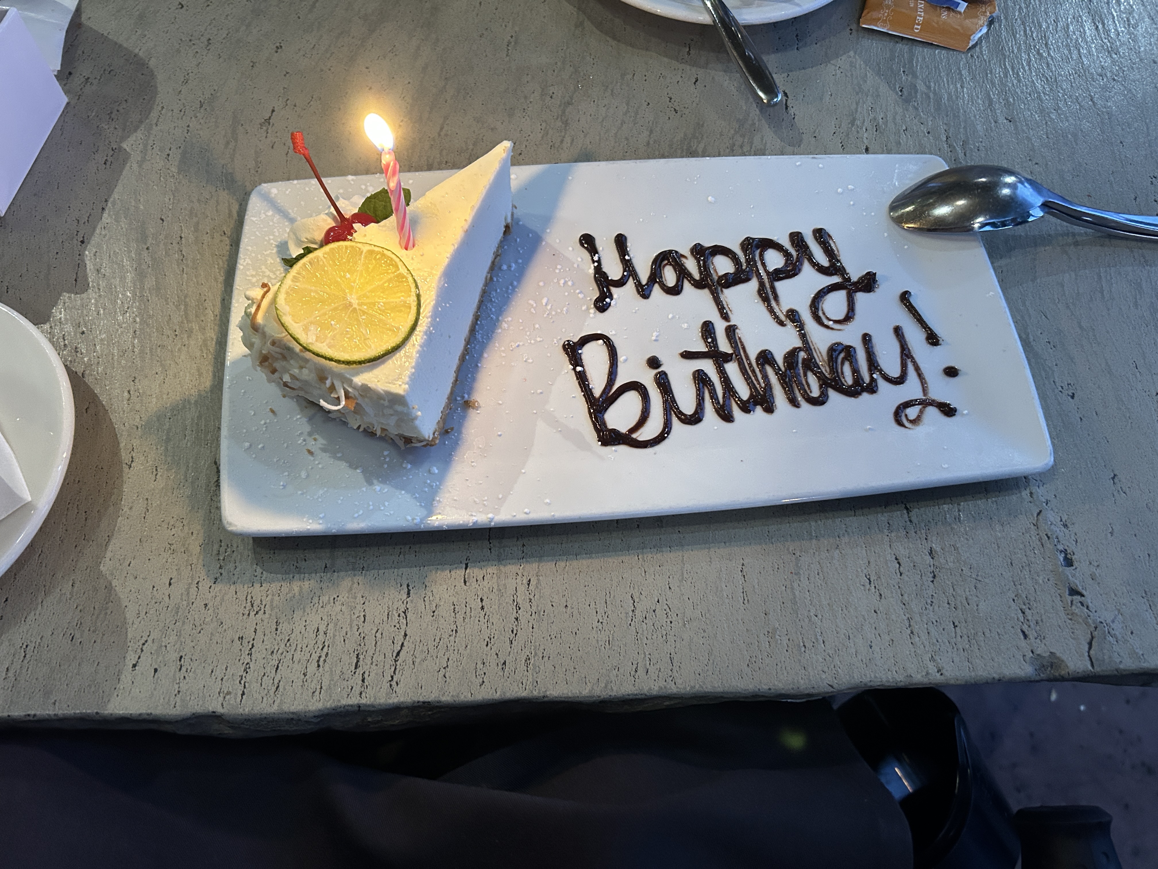 A slice of cheesecake with a lit pink candle sits beside “Happy Birthday!” written in chocolate.