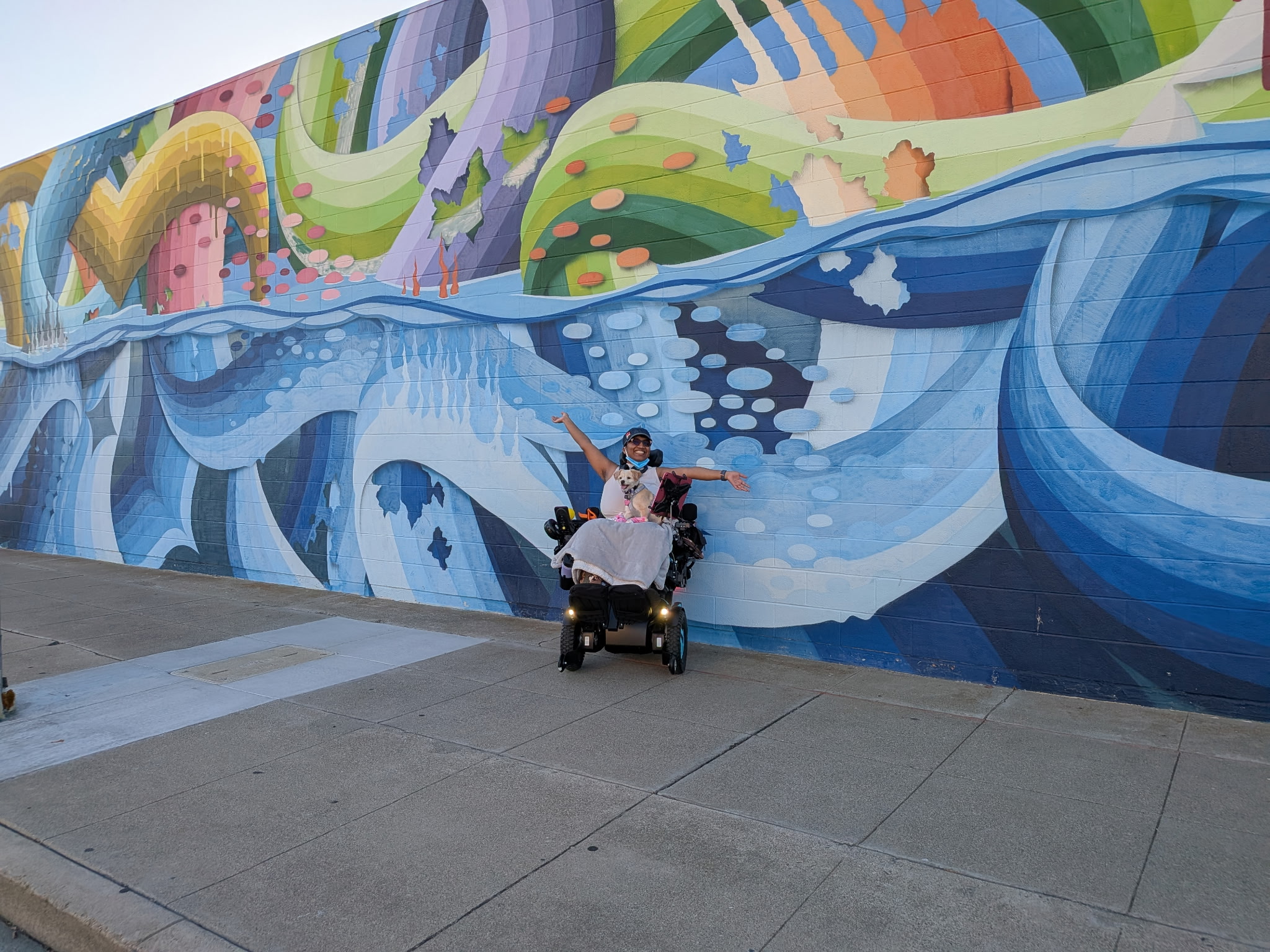 Harshini sits in her power wheelchair with Toffee against her chest and both arms raised in front of a large colorful mural.