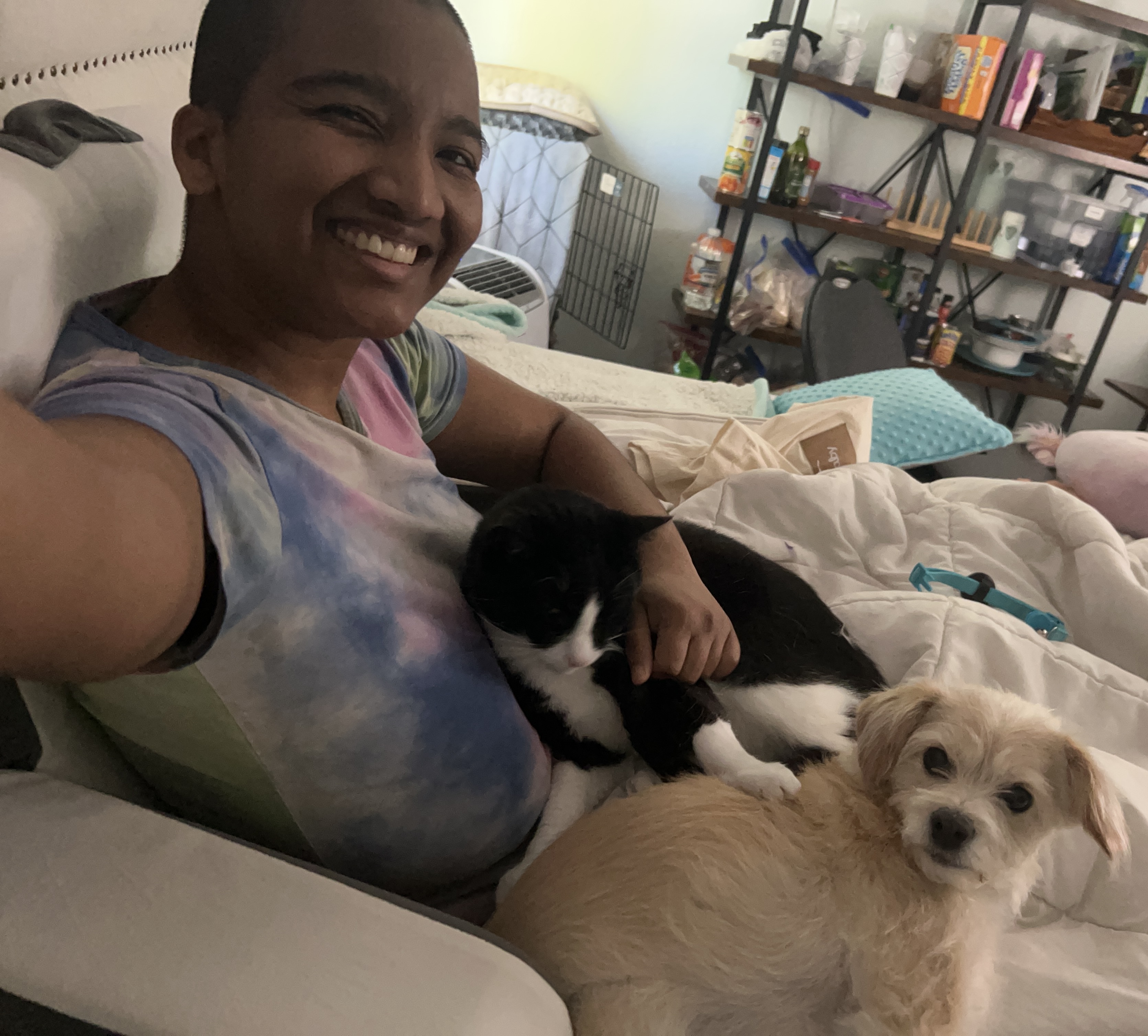 A smiling person sitting in bed with a black-and-white cat resting on their lap and a small cream-colored dog lying next to them.
