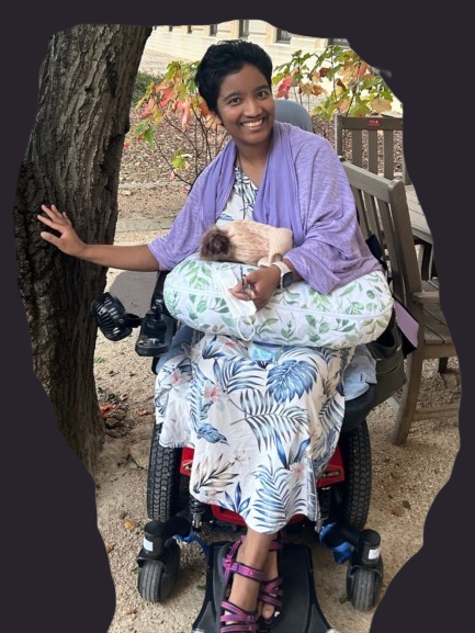 Harshini Busireddy smiling outdoors in a power wheelchair with a small animal resting on a pillow in her lap.