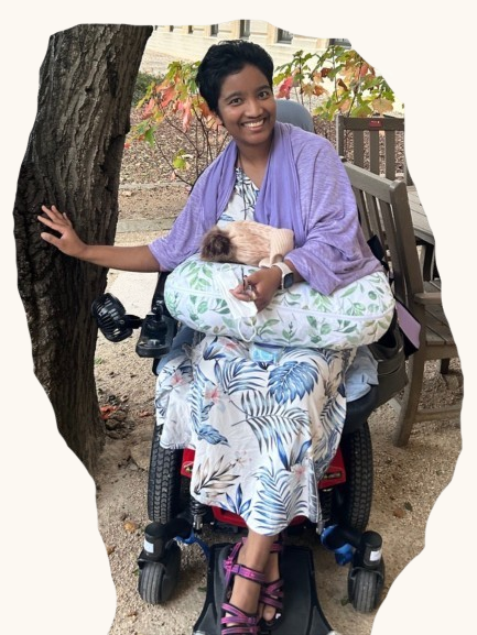 Harshini Busireddy smiling outdoors in a power wheelchair with a small animal resting on a pillow in her lap.