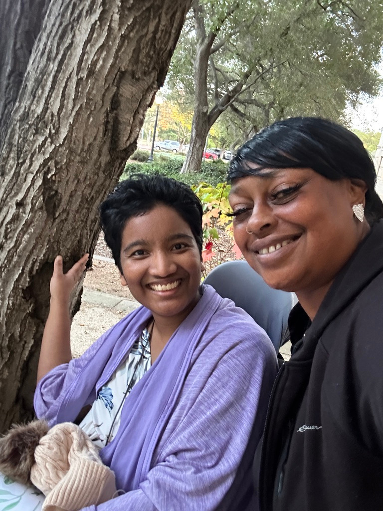 Harshini outdoors beside a tree with her care provider, both smiling softly.