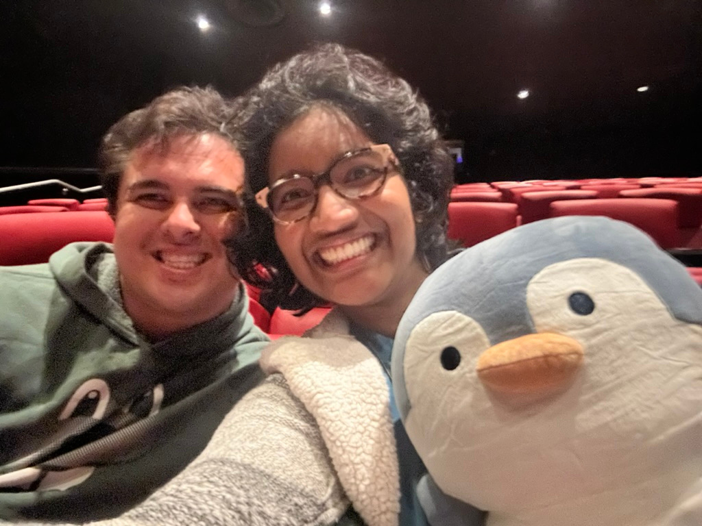 Harshini and her friend seated in a movie theater with a penguin plush, smiling together.