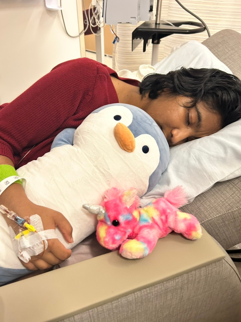 Harshini resting on a hospital chair, holding a penguin plush close, and a pink toy unicorn on the armrest.