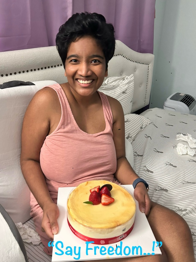 Harshini sitting on a bed, smiling while holding a cake topped with strawberries. Words 'Say Freedom..!' are marked on the image.