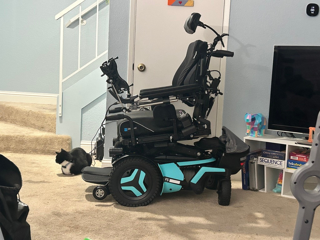 Harshini's custom power wheelchair parked in a living room, with her cat nearby.