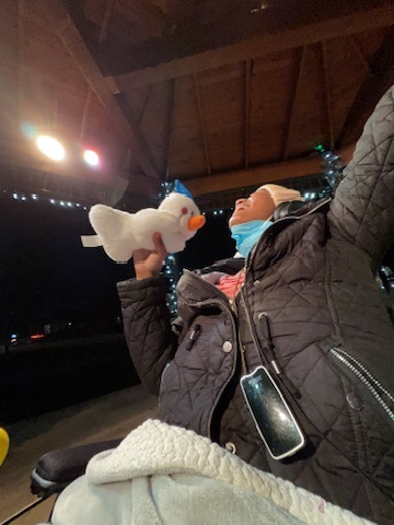 Harshini outdoors in a wheelchair at night, posing with a snowman plush as a mic with her arms out.